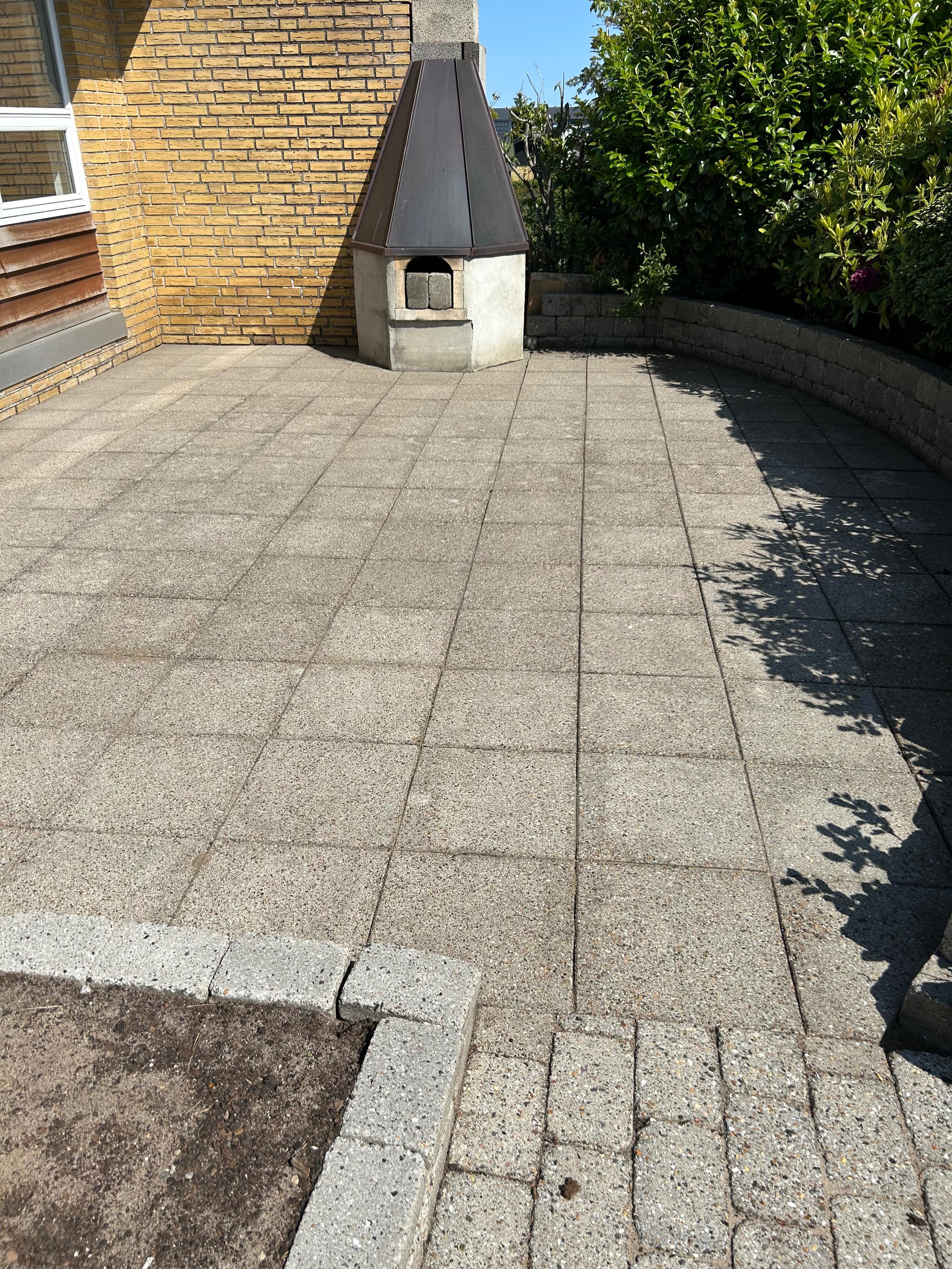En flisebelagt terrasse med en grill i baggrunden og grønne buske langs kanten.