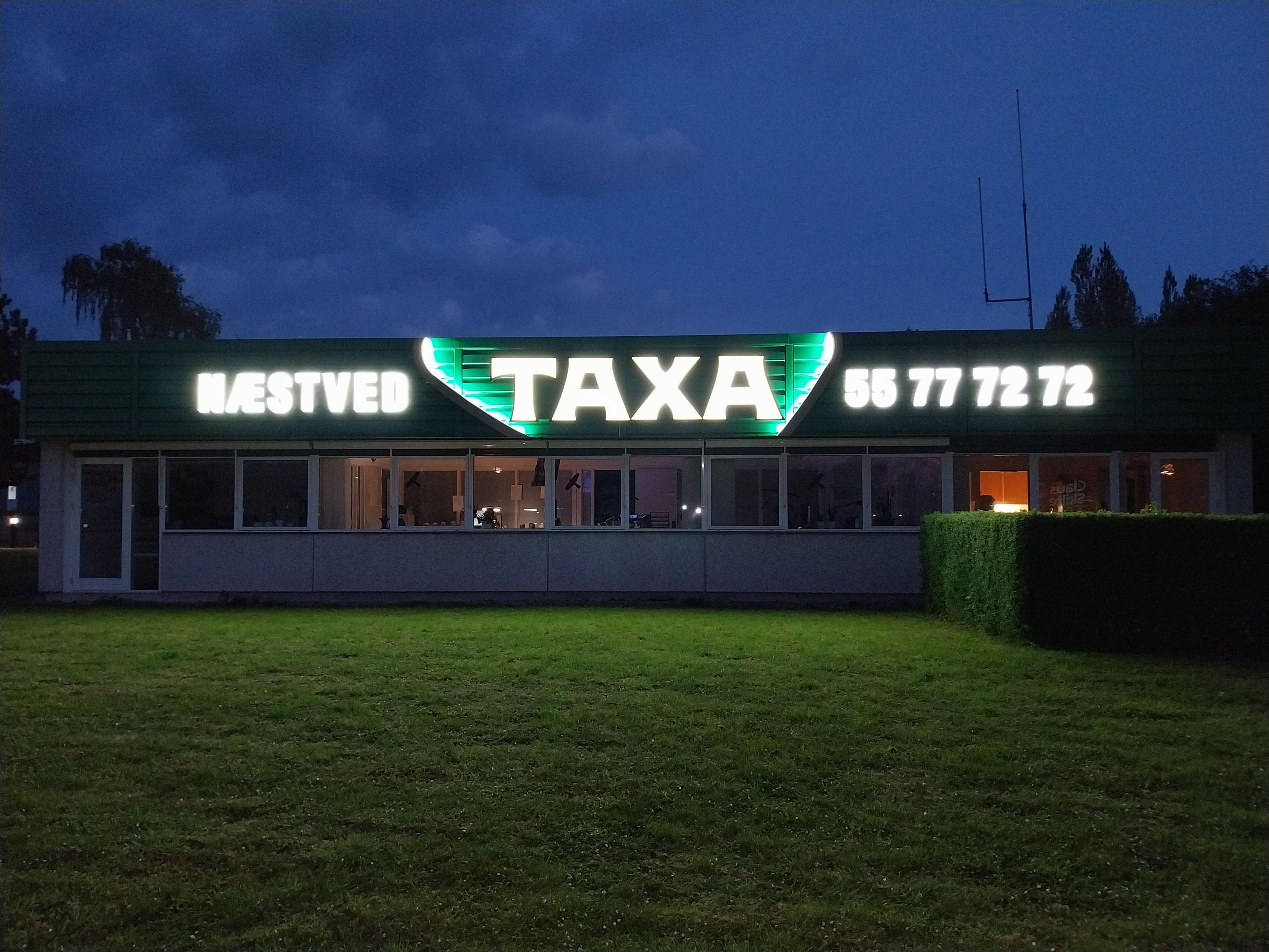 Taxa-skilt med lys i Bæstved, viser telefonnummeret 77 72 72 på en bygning om aftenen.