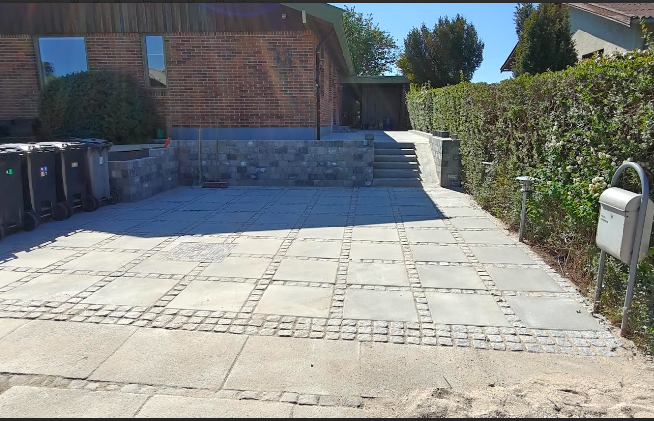 En indkørsel med fliser, affaldscontainere og en trappe, omgivet af buske og et hus i baggrunden.