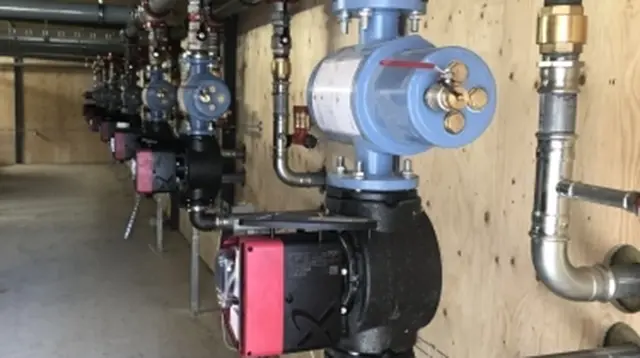 Rørinstallation med pumper og ventiler i et teknisk rum med trævægge.