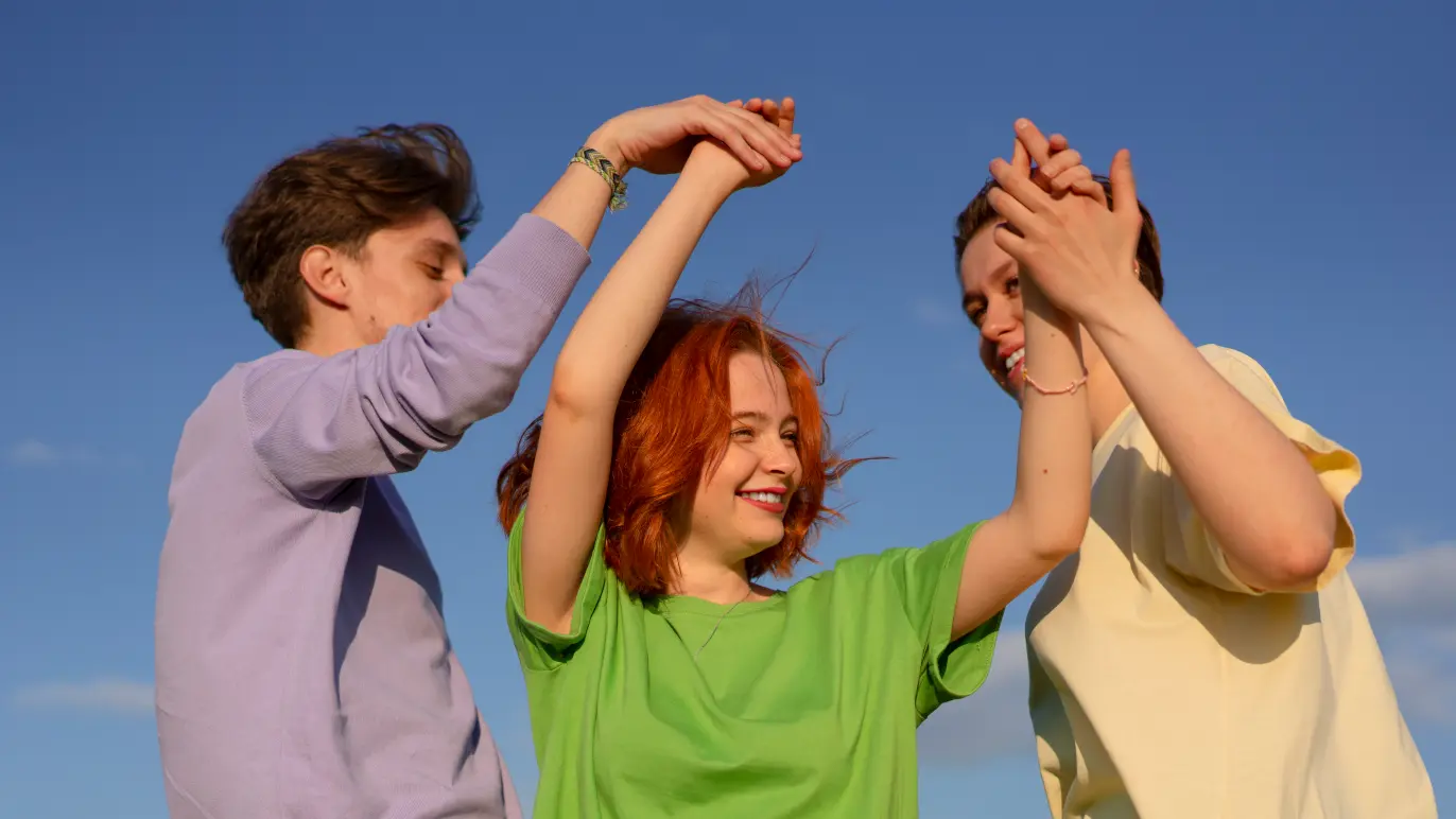 Tre unge mennesker danser og holder hinandens hænder under en blå himmel med skyer.