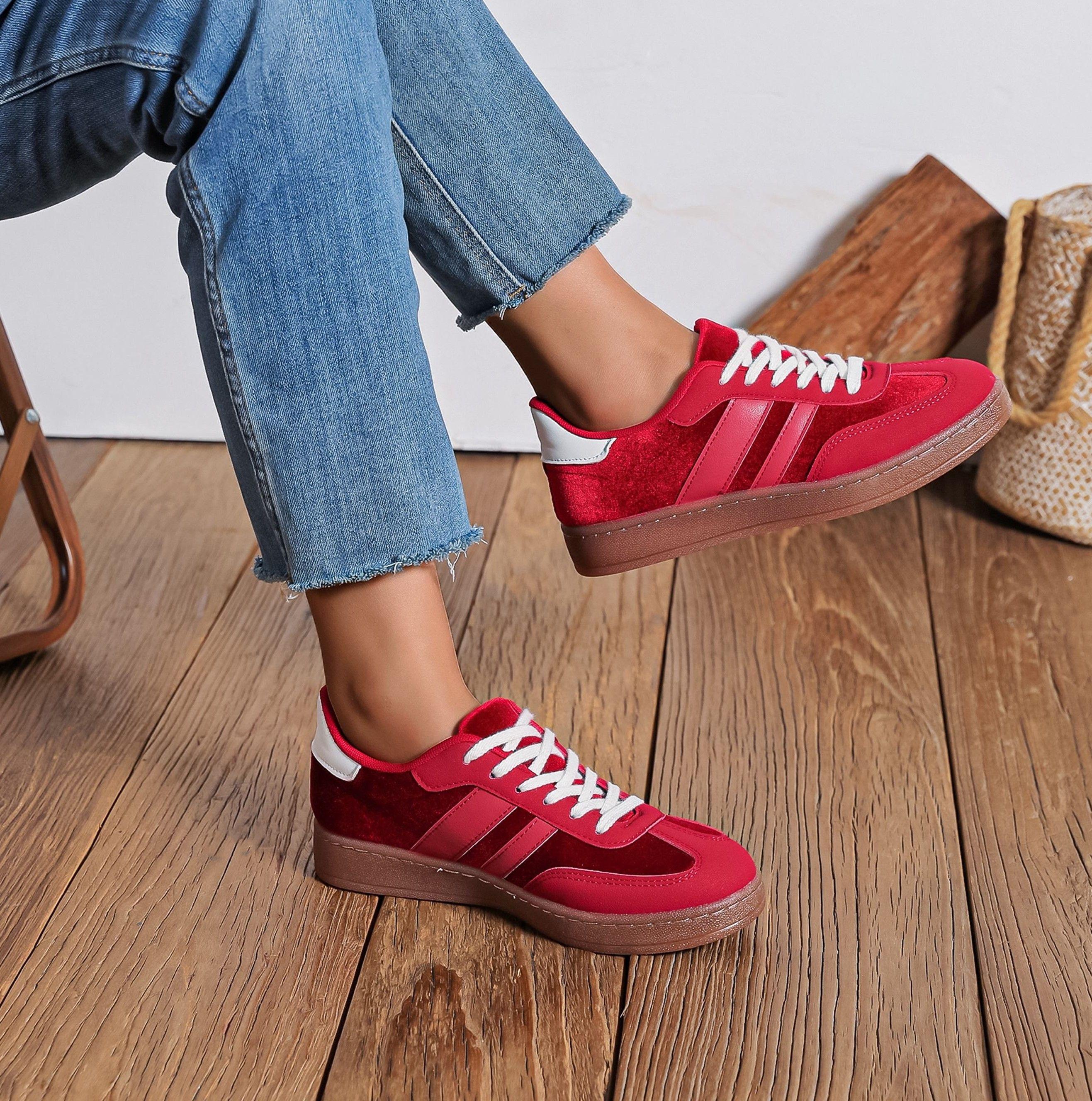 Rød sneakers med hvid snørebånd, båret af en person i jeans, står på et trægulv.