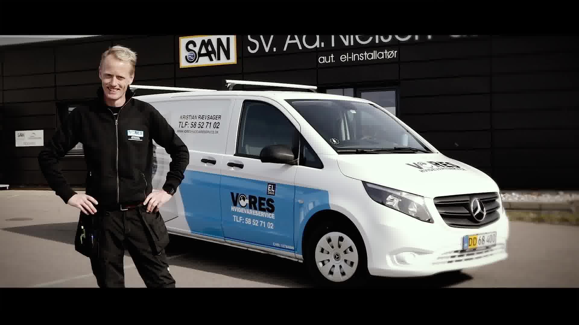 En smilende håndværker står ved siden af en hvid servicevogn med blå detaljer foran en virksomhed.