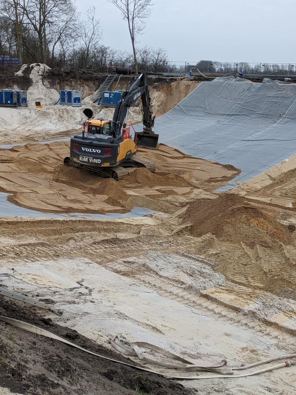 En gravemaskine arbejder i en sandgrav med sand og en træplante i baggrunden.