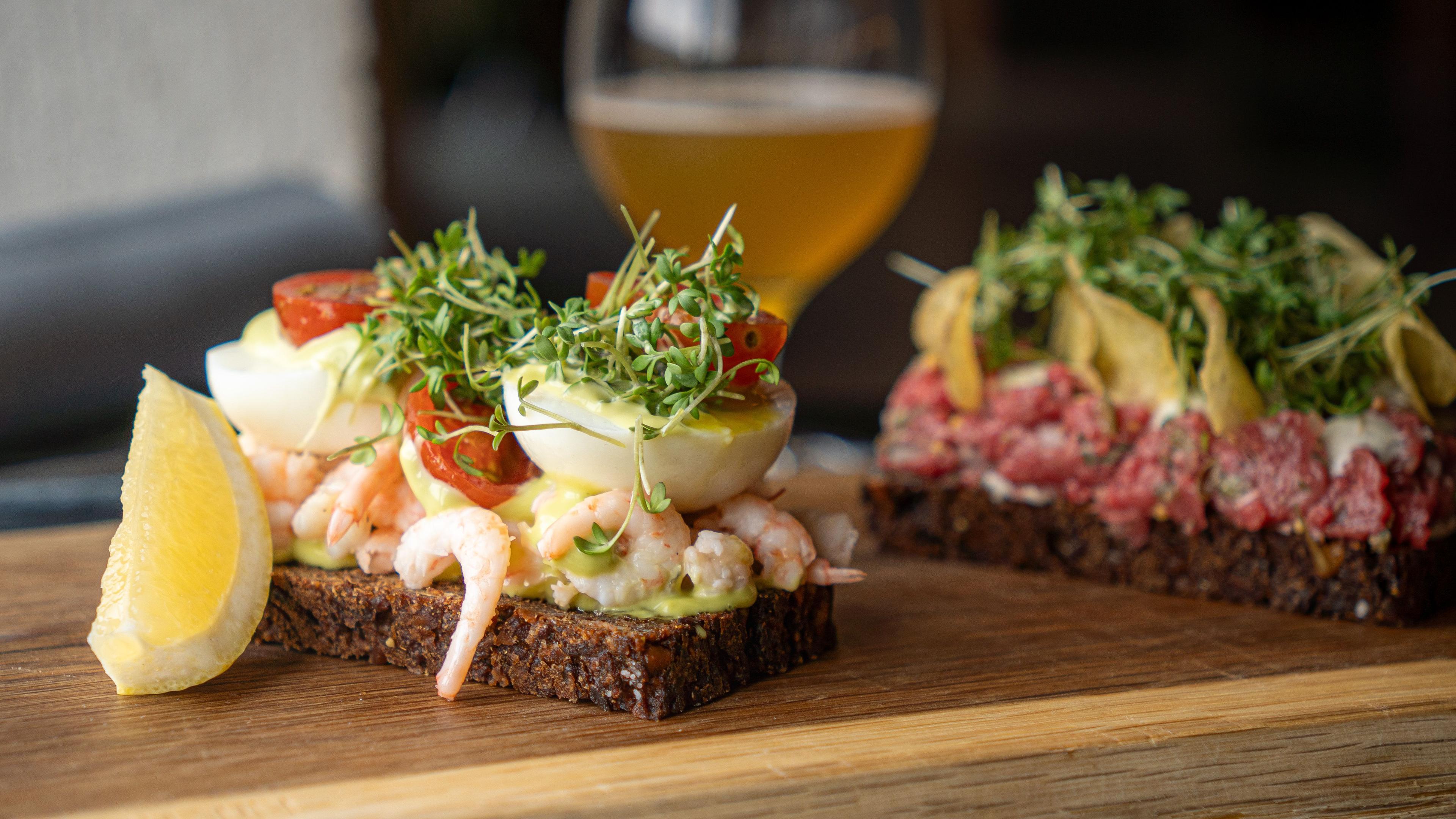Toasted rugbrød med rejer, æg og tomater, serveret med citron og en øl i baggrunden.