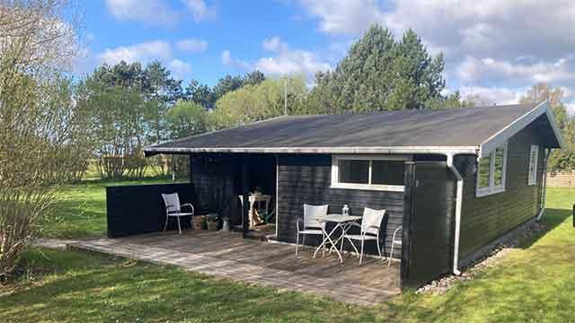 Et sort sommerhus med terrasse, omgivet af grønne træer og græs, under en blå himmel med skyer.