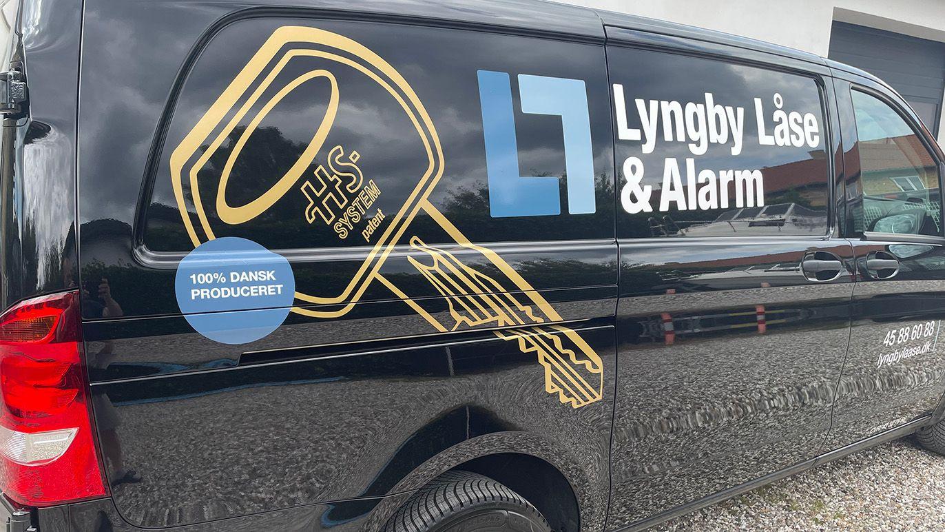 Sort varevogn med logo for Lyngby Låse & Alarm og en stor nøgleillustration på siden.