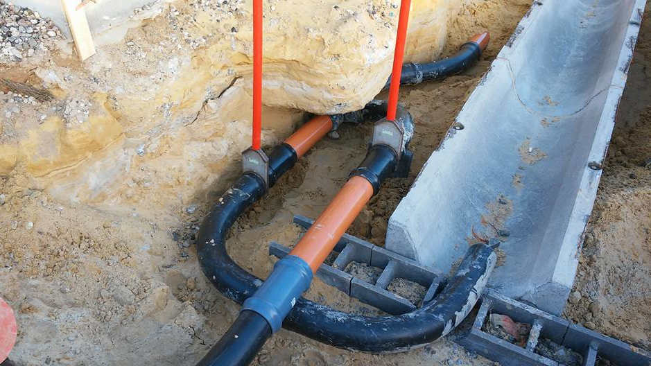 Rørinstallation i en byggegrav med sorte og orange rør samt betonkanal i baggrunden.