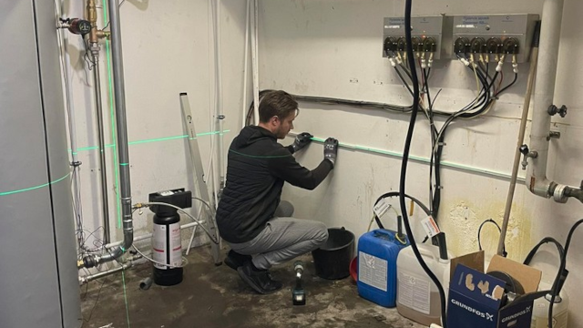 En mand arbejder med rørinstallation i et kælderrum, mens han bruger en laserlinje til præcision.