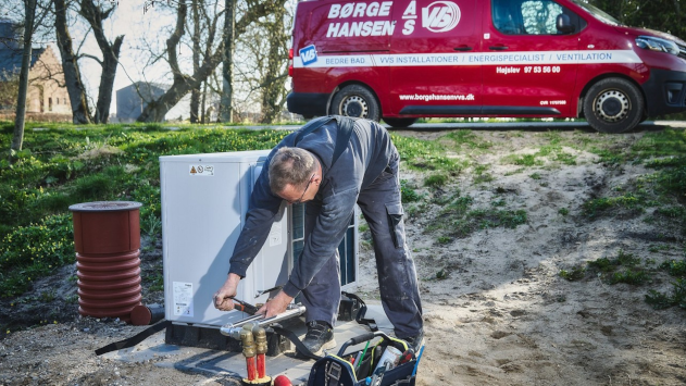 En håndværker arbejder på en varmepumpe ved en rød servicevogn fra Borge Hansen.