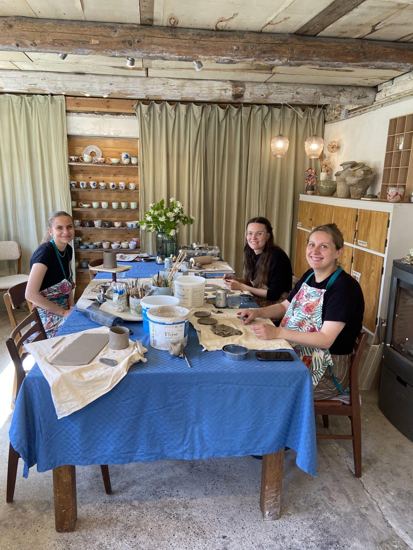 Tre personer sidder ved et bord i et lyst rum, mens de arbejder med keramik og kunsthåndværk.