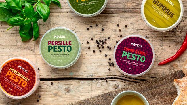 Forskellige pestoer og hummus i skåle på et træbord med friske krydderurter og peberkorn.