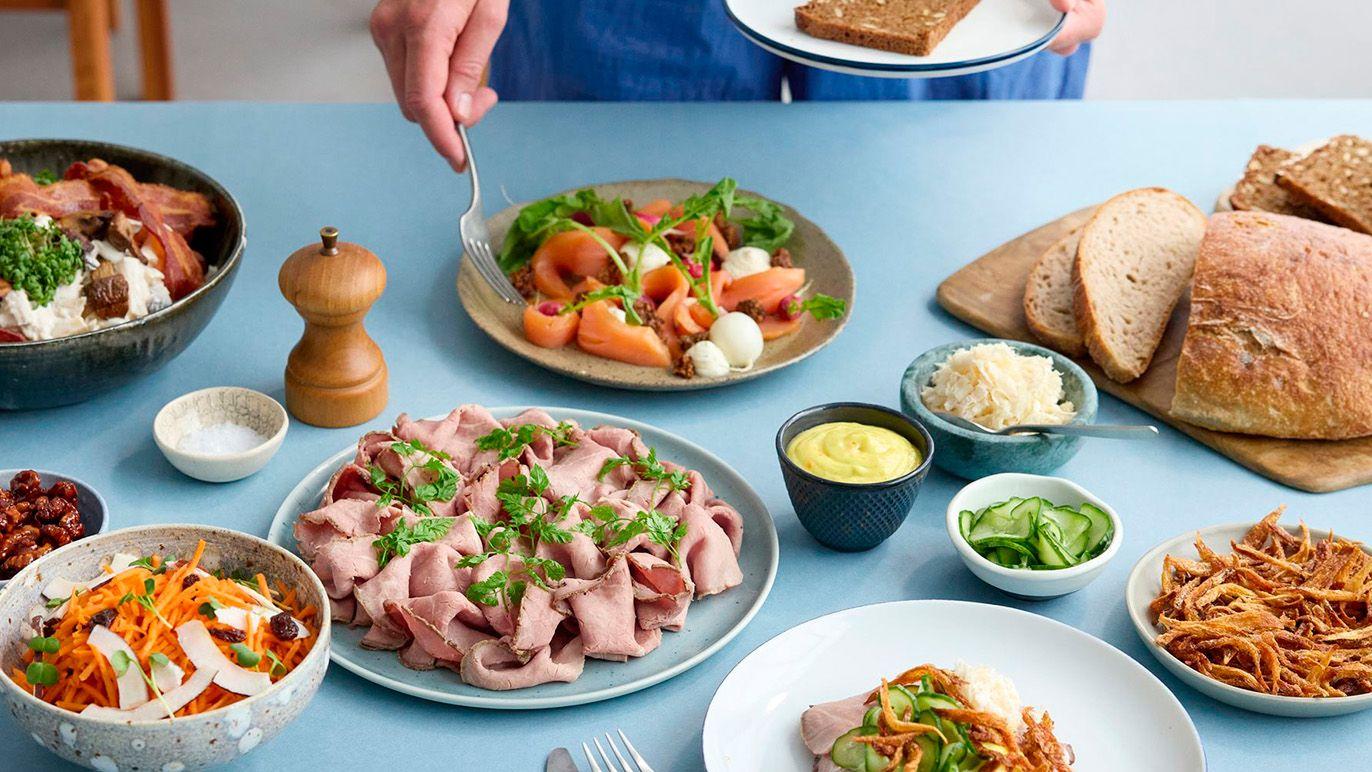 En person serverer mad ved et bord med forskellige retter, herunder pålæg, salat, brød og tilbehør.