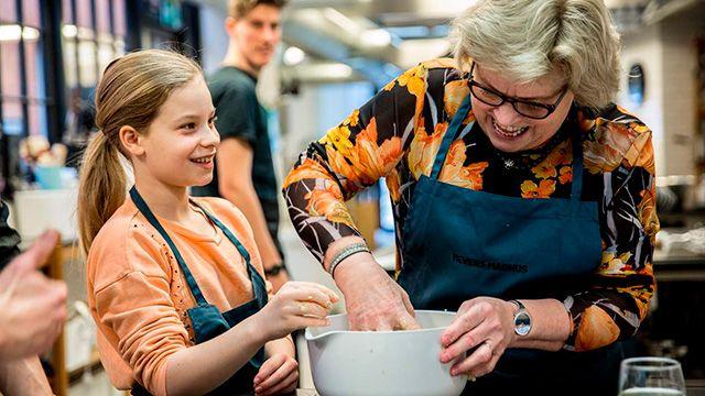 En glad pige og en ældre kvinde laver mad sammen i et køkken, begge iført forklæder.