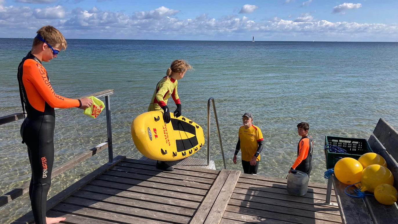 Børn i våddragter leger ved stranden, en dreng bærer en surfeboard, mens andre står i vandet.