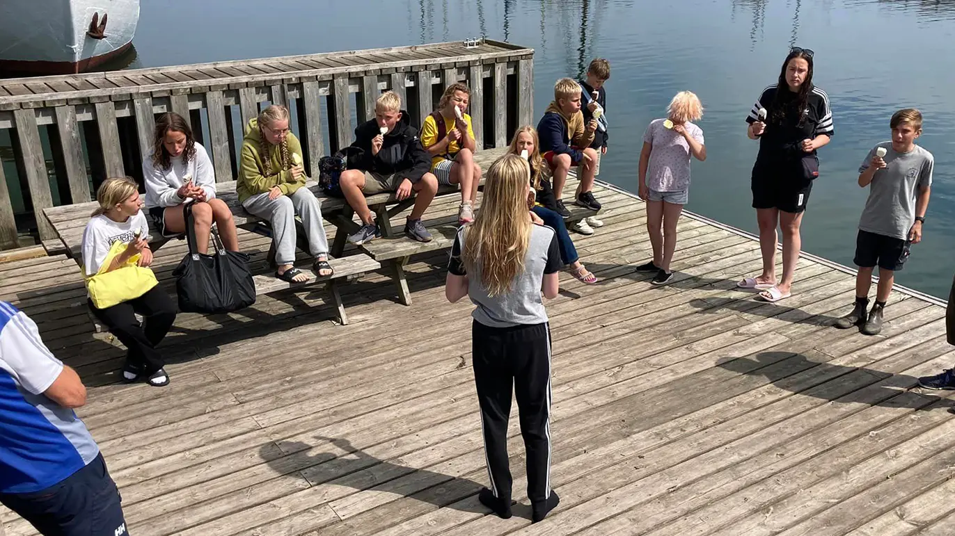 Gruppe af børn og unge, der sidder og står på en træbro ved vandet, mens de snakker og spiser.