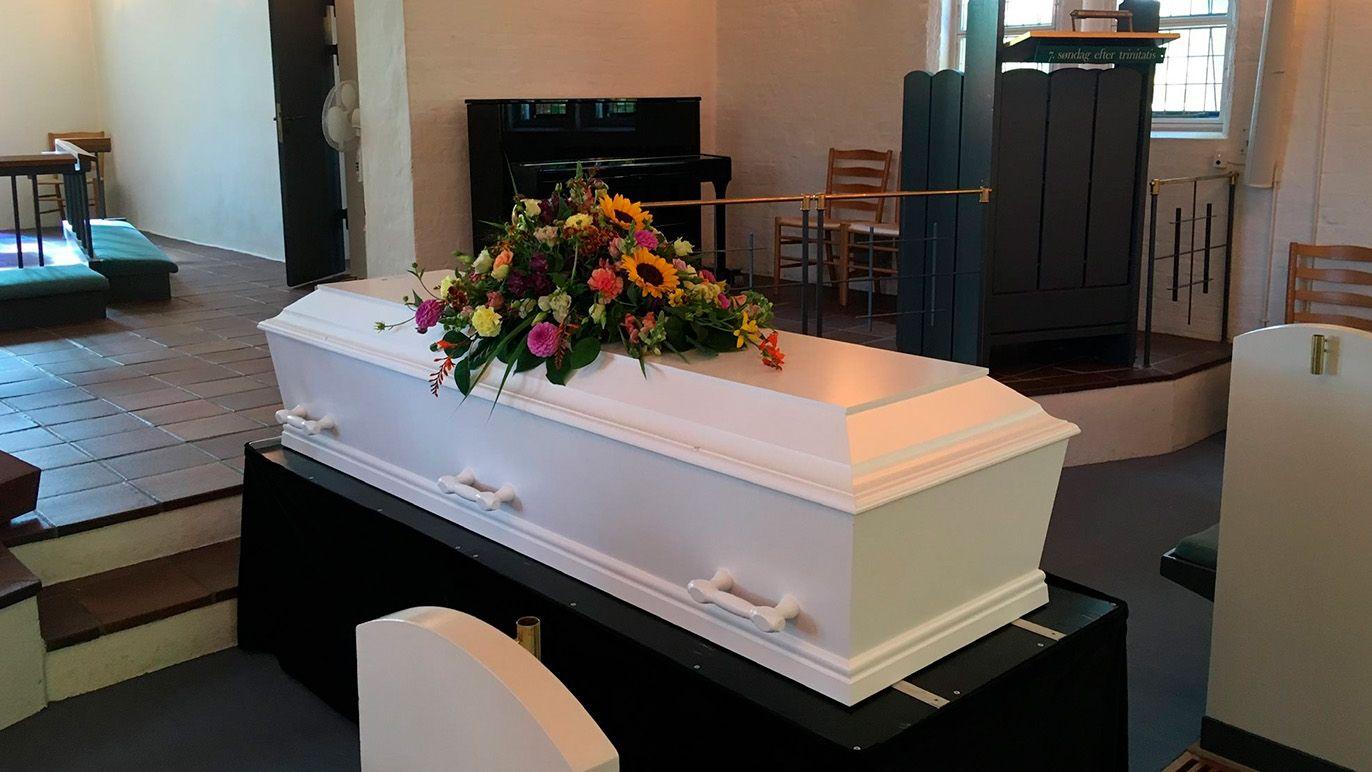 En hvid kiste med blomsterarrangement står i et rum med stole og et klaver i baggrunden.
