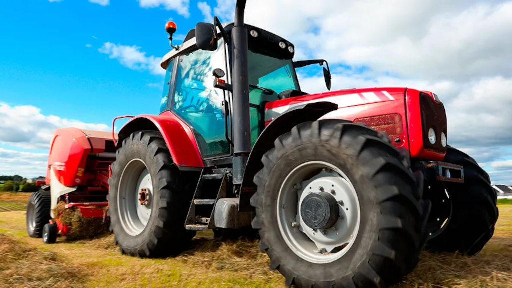 Rød traktor med store dæk arbejder på en mark under en blå himmel med skyer.