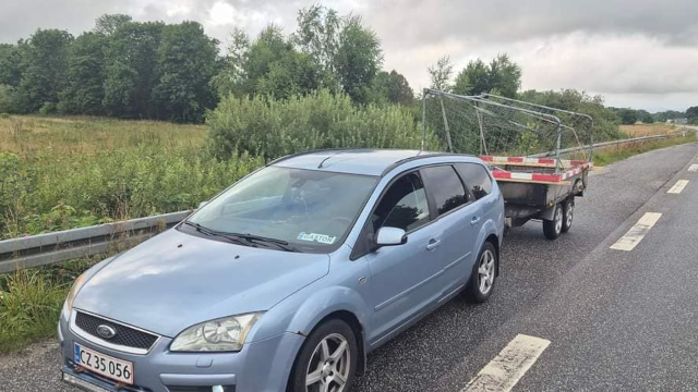 En blå Ford stationcar trækker en trailer med en metalramme langs en landevej omgivet af grønne træer.