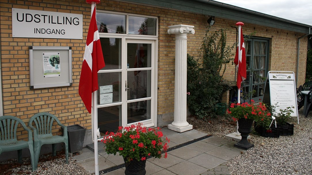Indgangen til en udstillingsbygning med danske flag og blomsterkummer foran.