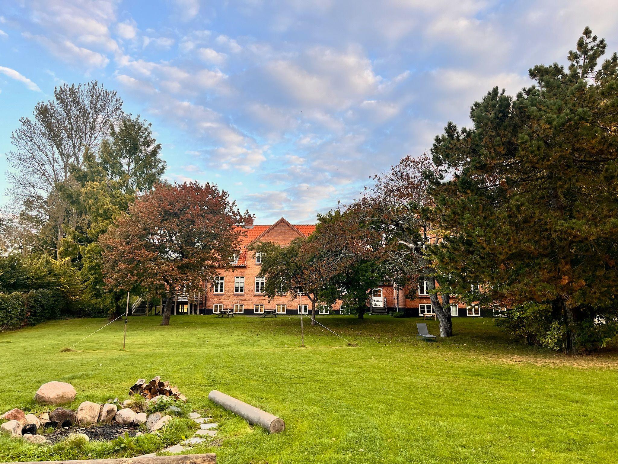 En grøn græsplæne med træer og en bygning i baggrunden under en skyet himmel.