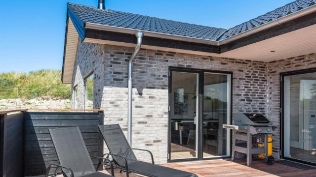 Moderne hus med grå mursten, terrasse, liggestole og grill under en klar blå himmel.