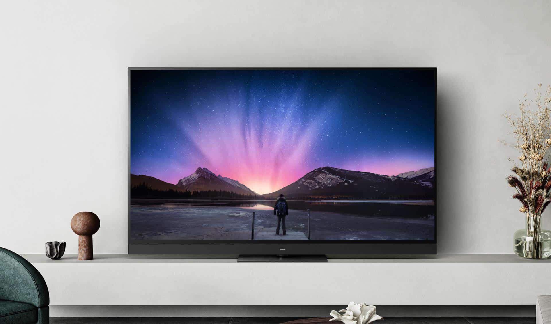 En moderne tv på en væg med en person, der står ved en sø og ser på en farverig solnedgang over bjergene.
