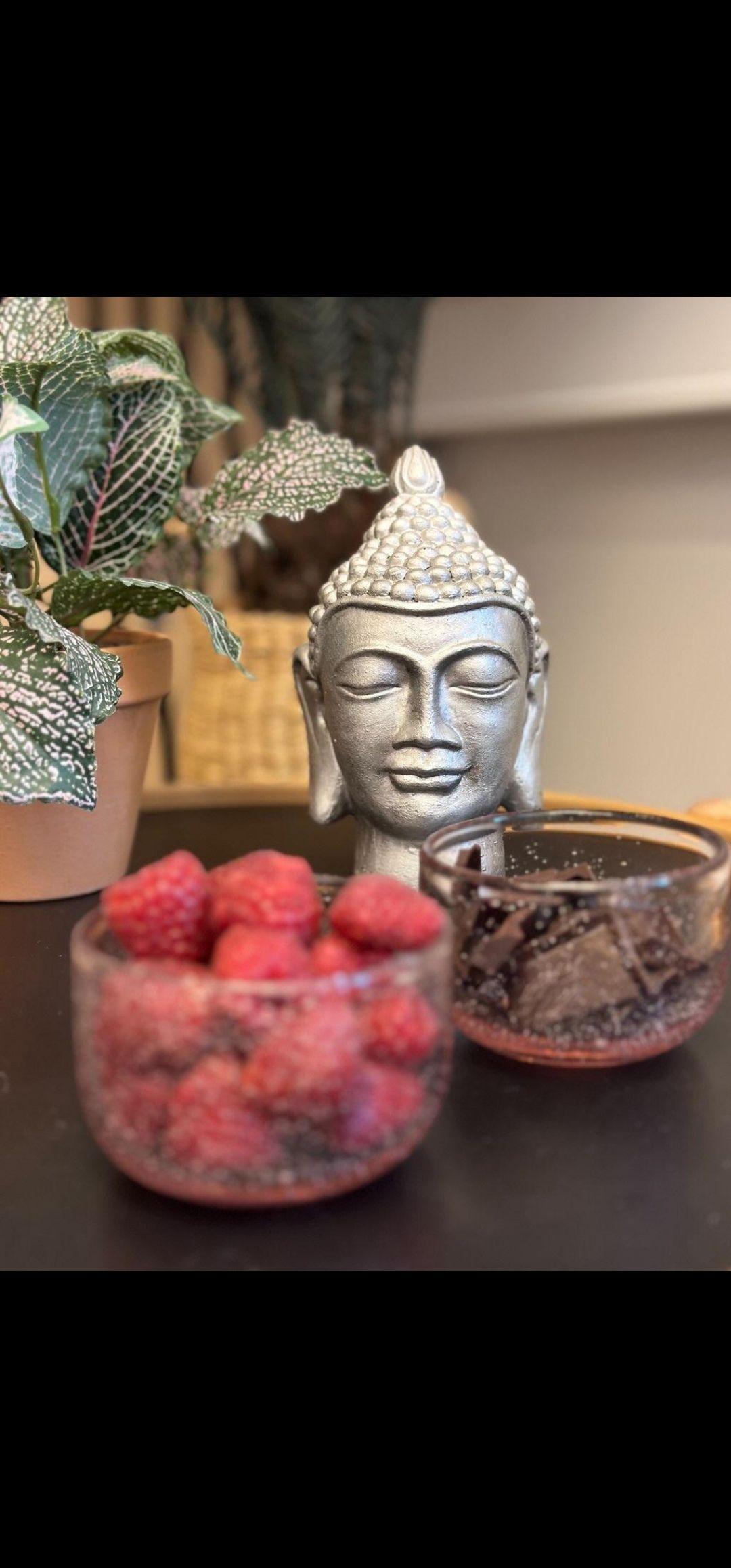 Buddha statue med lukkede øjne, skål med hindbær og en skål med chokolade på et bord.