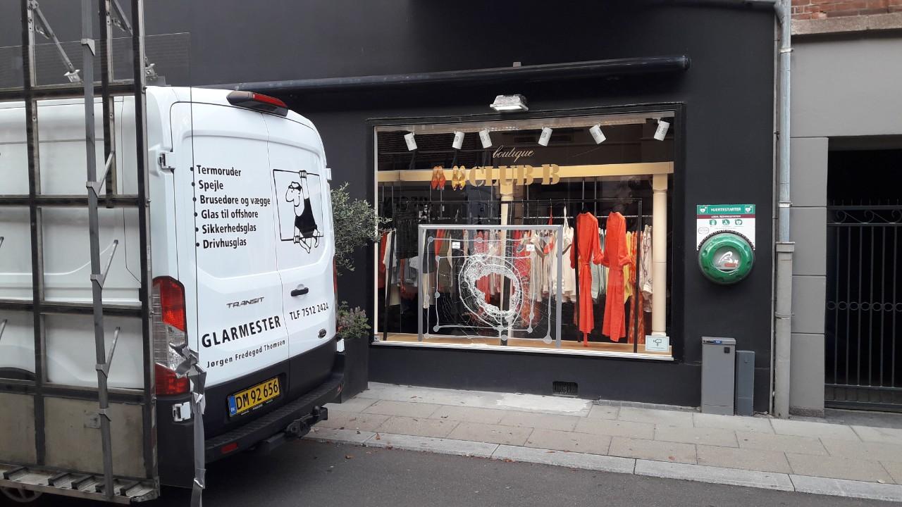 Butiksvindue med orange tøj og en varevogn parkeret foran, der viser tekst og logo.