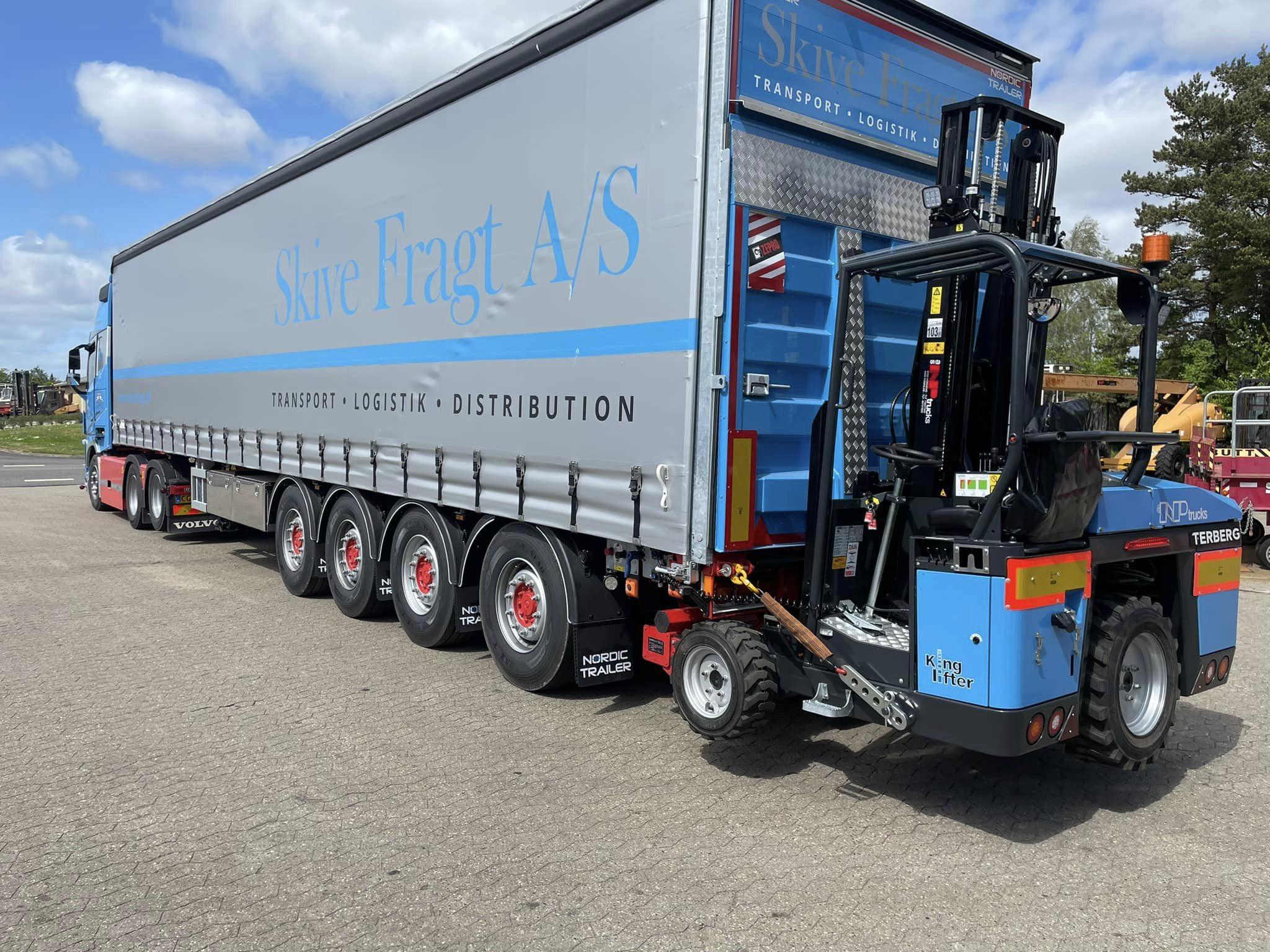 En stor lastbil med en blå og grå trailer, der står parkeret på en grusvej under en blå himmel.