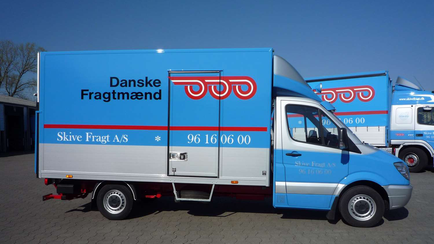 Blå fragtbil fra Danske Fragtmænd med logo og kontaktinformation på siden, parkeret under en klar blå himmel.