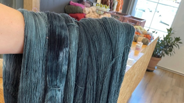 En hånd holder en bundt blågrønt garn foran en butik med farverigt garn og strikkeudstyr i baggrunden.