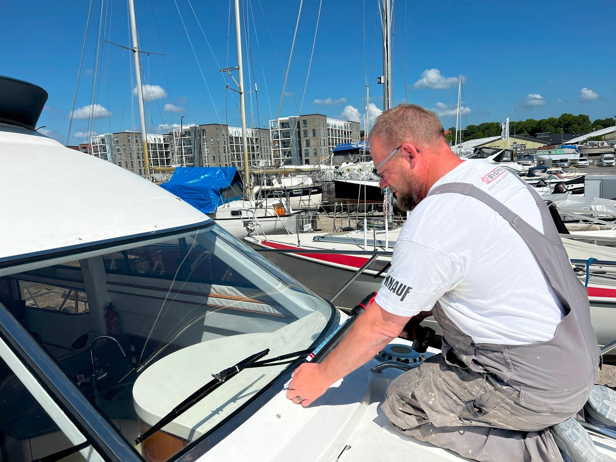 Mand arbejder på en båd i havnen, omgivet af både og bygninger under en klar blå himmel.