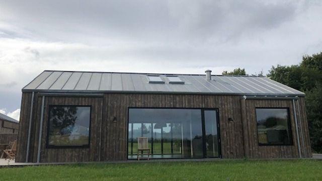Moderne træhus med store vinduer og metaltag, beliggende i et grønt landskab under en skyet himmel.