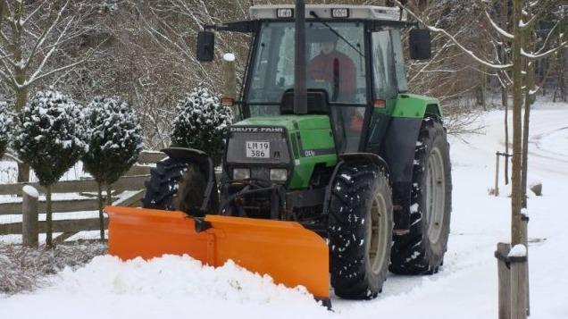 Traktor med orange sneplov, der rydder sne på en vintervej omgivet af træer og hegn.