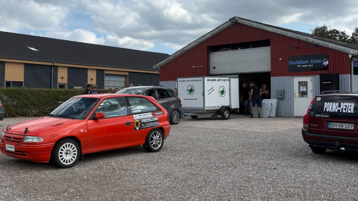 Rød bil parkeret foran en garage med en trailer og personer, der arbejder i baggrunden.