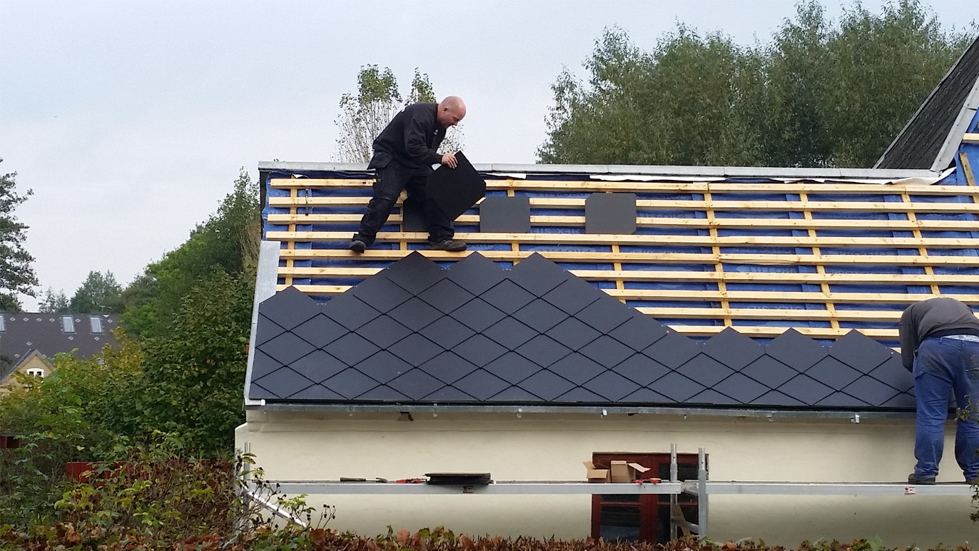 To personer arbejder på at lægge tagplader på et hus med et skråt tag i et grønt område.