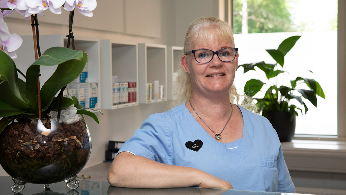En smilende kvinde i blå uniform står ved en reception med planter og blomster i baggrunden.