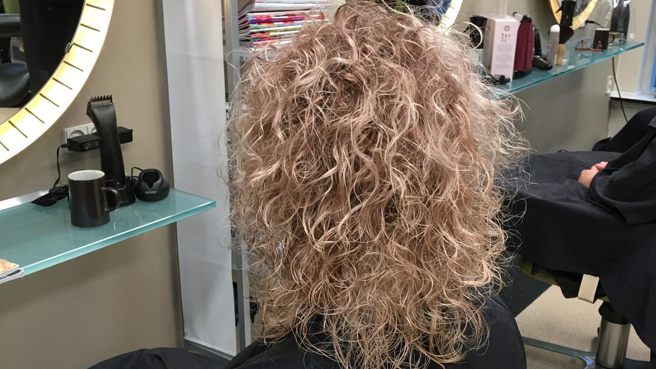 En person med krøllet, blond hår sidder i en frisørsalon foran et spejl.