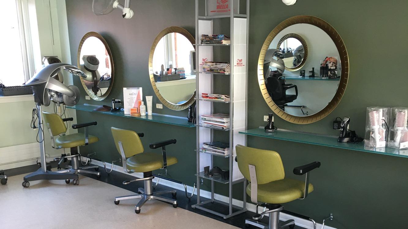 Frisørsalon med grøn væg, spejle, frisørstole og hylder med skønhedsprodukter.