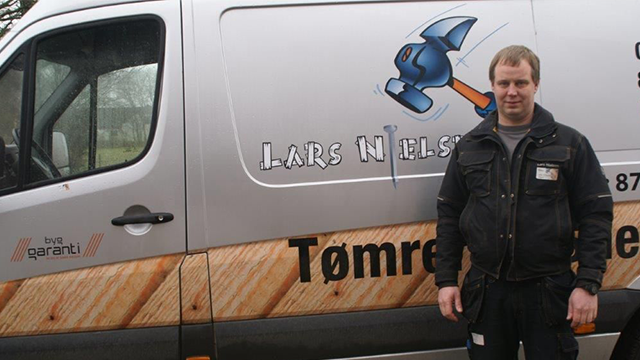 En håndværker står ved siden af en varevogn med teksten "Lars Nielsen Tømrer" og et billede af en hammer.
