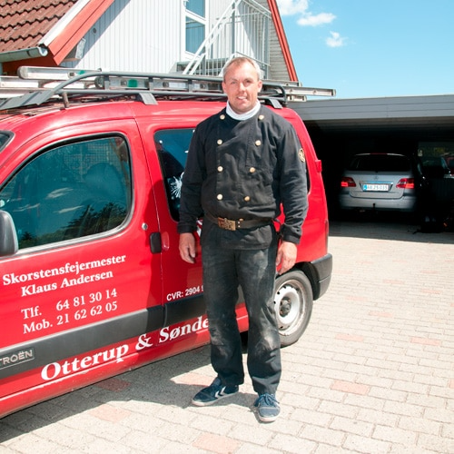 En skorstensfejer står foran sin røde bil med logo, klar til arbejde i en garageopkørsel.
