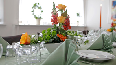 Smukt dækket bord med blomsterarrangement, grønne servietter og glas i et lyst rum.