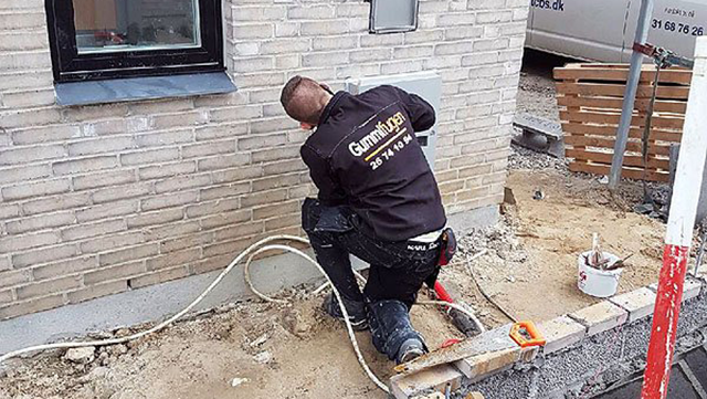 En håndværker arbejder på en mur med værktøj og ledninger ved en bygning.