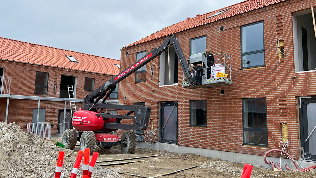 Byggeri med en lift, der maler vinduer på en murstensbygning med rød tag. Arbejdere i gang med renovering.