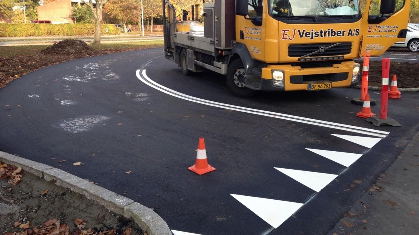 Lastbil fra Vejstriber A/S arbejder på at male vejstriber på en ny asfalteret vejkurve med advarselstriangler.