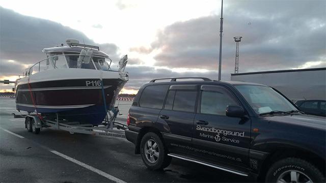 En bil trækker en båd på en trailer ved havnen under en skyet himmel.