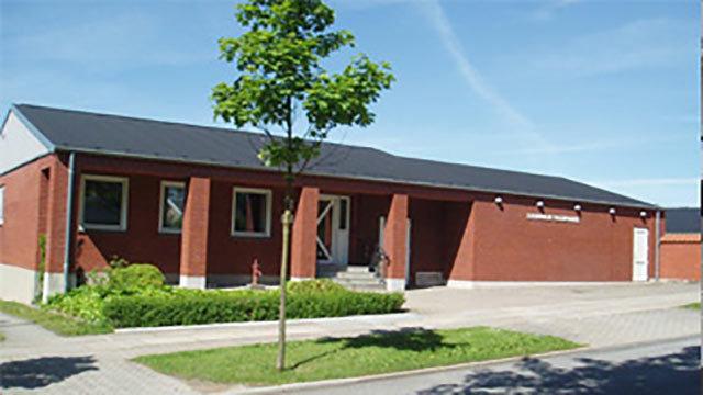 Rød murstensbygning med fladt tag og grønne buske foran, beliggende på en stille gade.