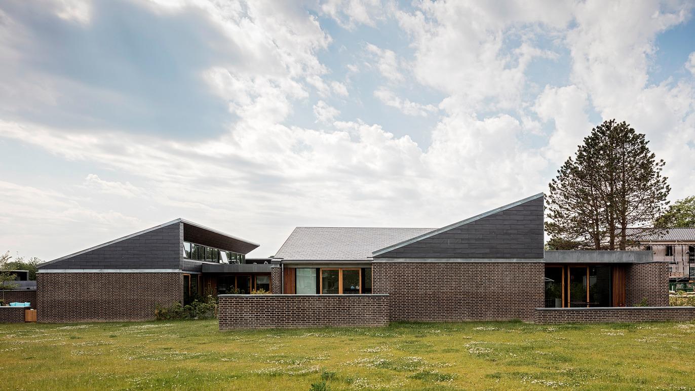 Moderne hus med skrå tag og store vinduer, omgivet af grøn græsplæne og træer under en skyet himmel.
