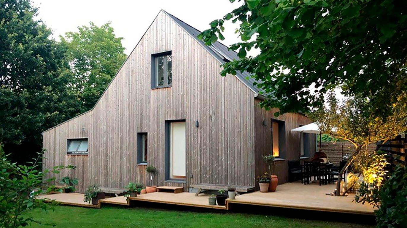 Moderne træhus med skrå tag, terrasse og grøn have, omgivet af træer og planter.